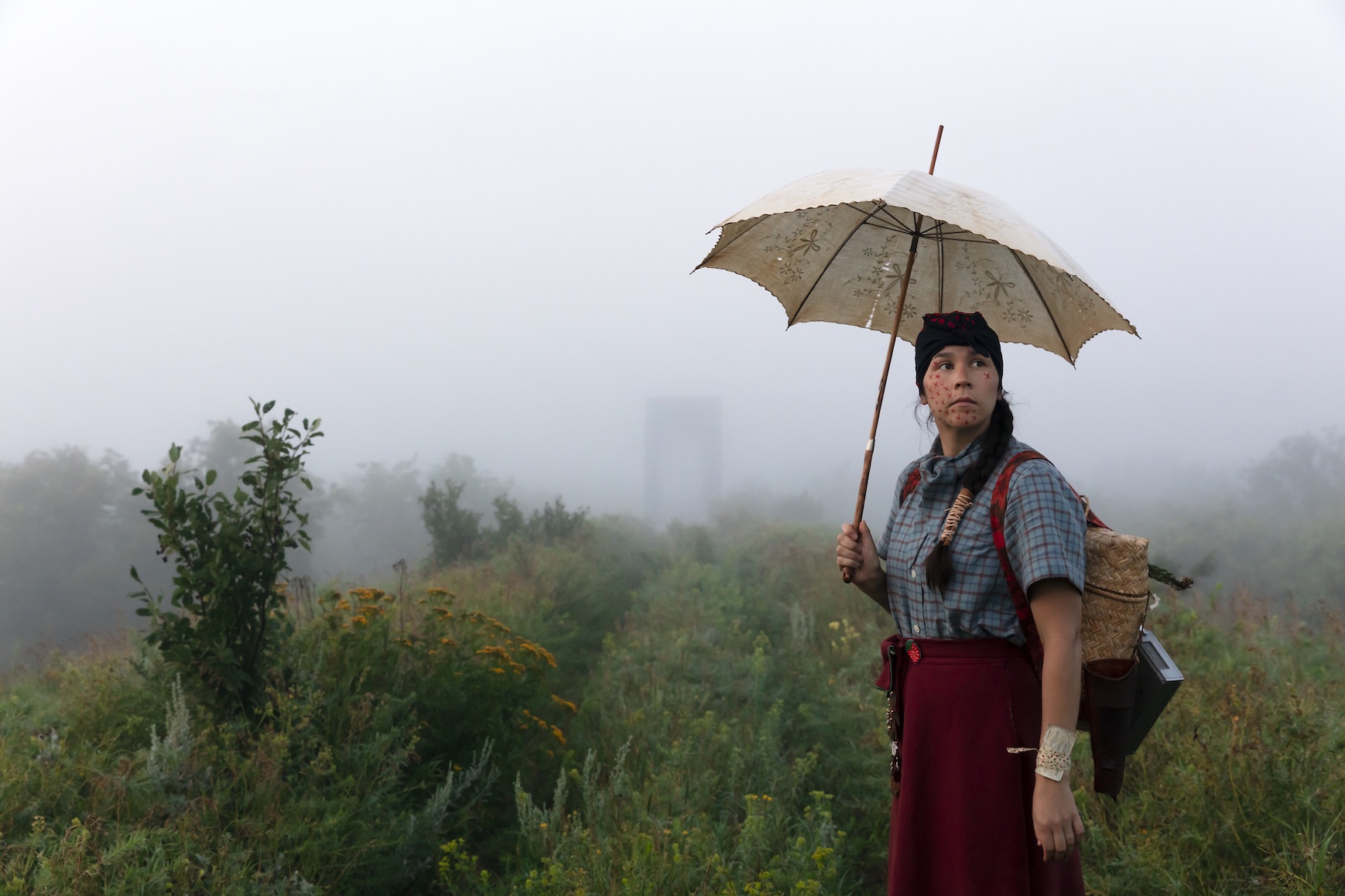 Self portrait of artist Meryl McMaster standing in a field in the fog with an umbrella