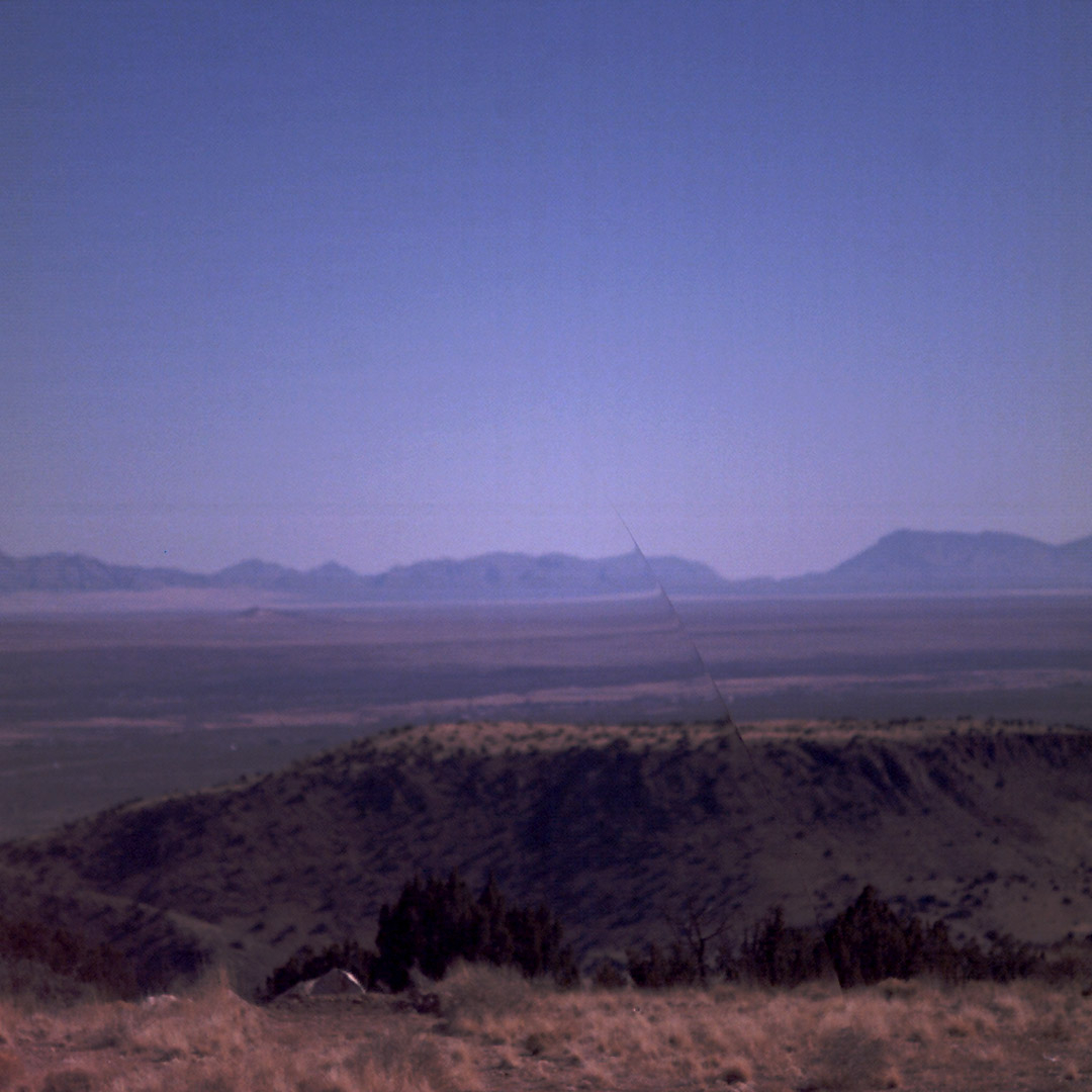 A square image show the landscape of the American West. Hills are seen in the background against a deep purple sky with dry grassland and brush in the foreground.