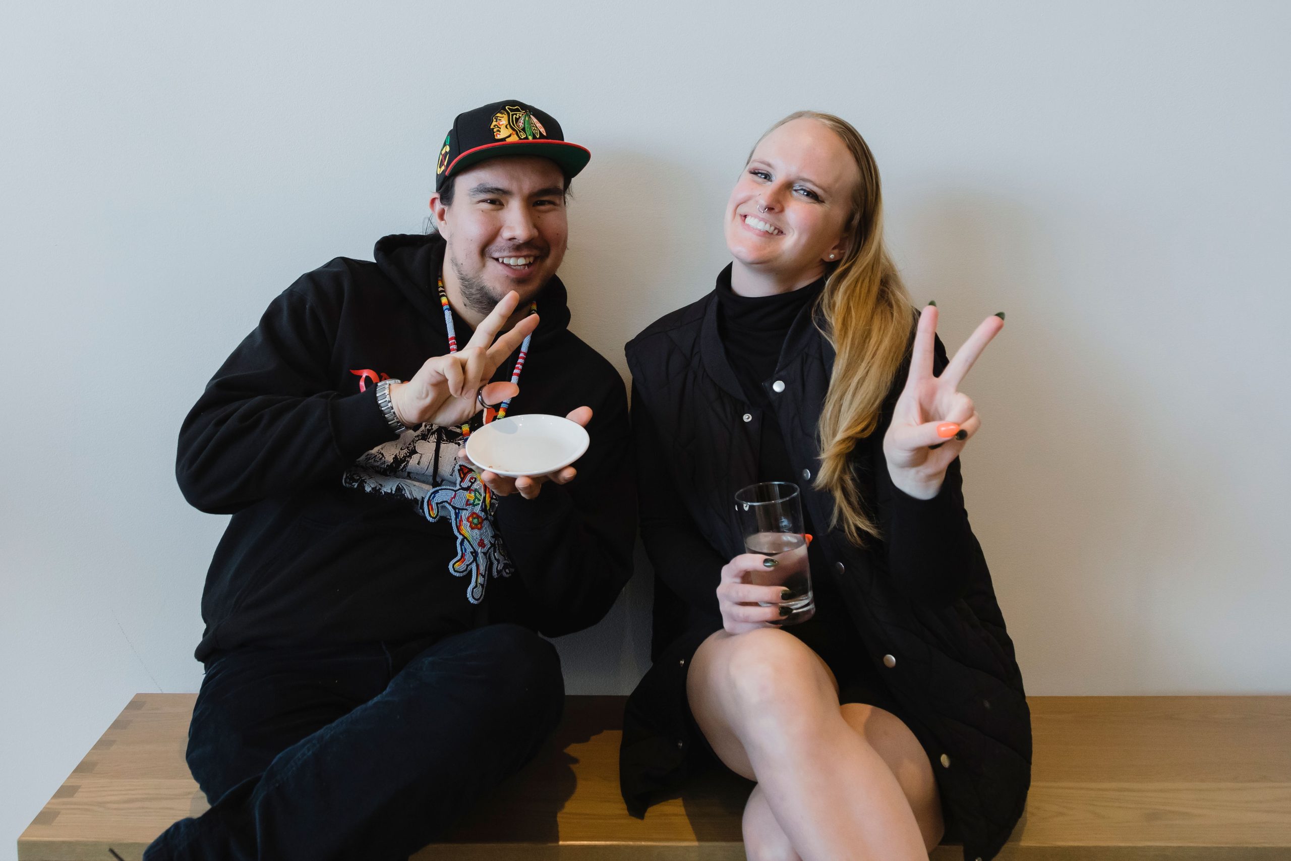 Two people sit on a bench together at Remai Modern, holding up peace signs to the camera.